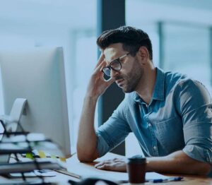 Frustrated man in a denim shirt sits at a desk, hand resting on his forehead while looking at a computer screen.