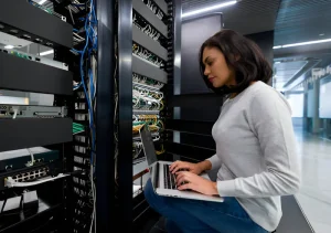 Woman seated by server racks in a data center, typing on a laptop while network cables and equipment loom around her.
