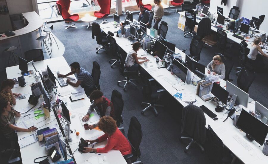 Open-plan office with long desks and multiple computer workstations; employees work at their computers amid large windows and scattered red lounge chairs in the background.