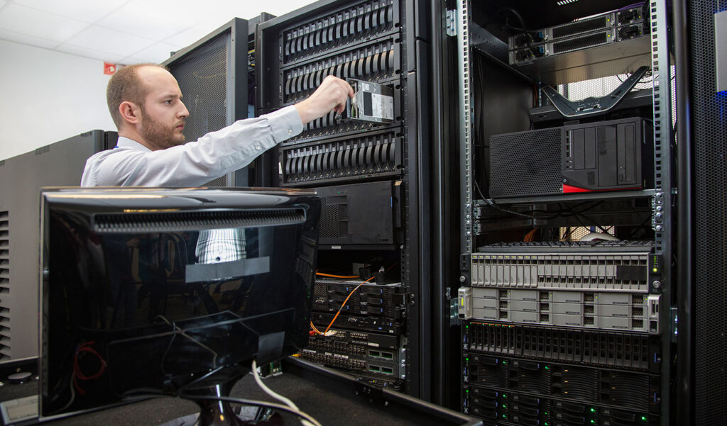 IT technician in a data center inserts a hard drive into a server rack among multiple stacked servers and networking gear.