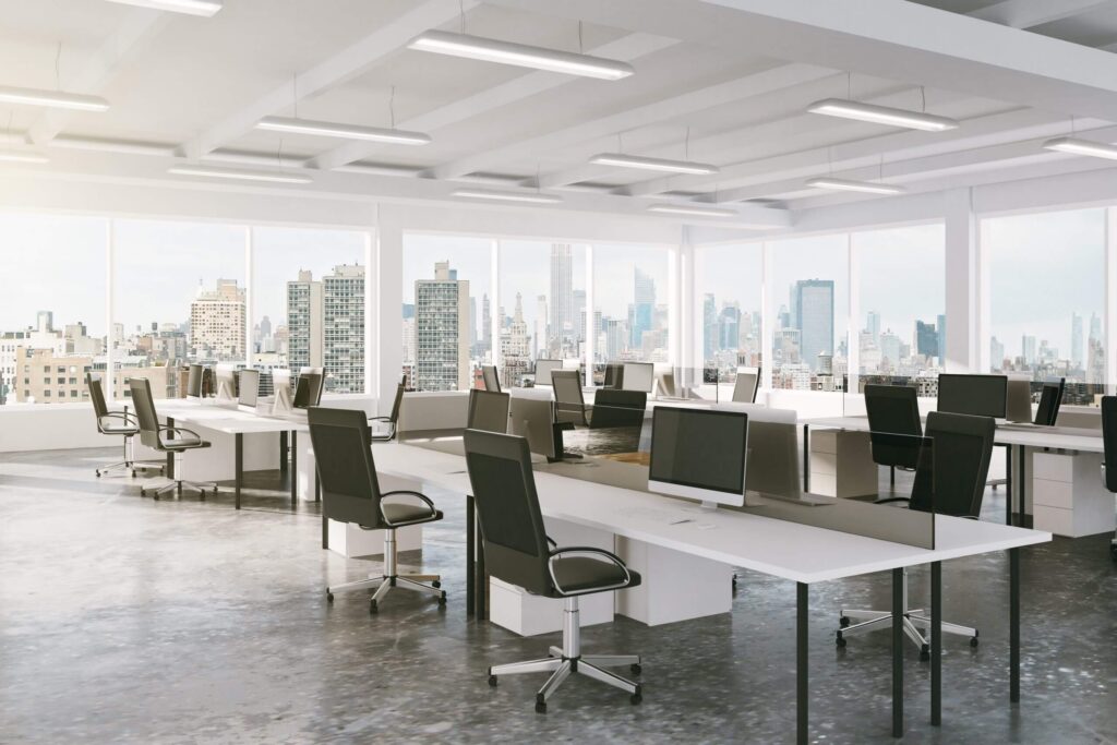 Open-plan office with rows of desks, ergonomic chairs, and multiple computer monitors, overlooking a city skyline through large windows.