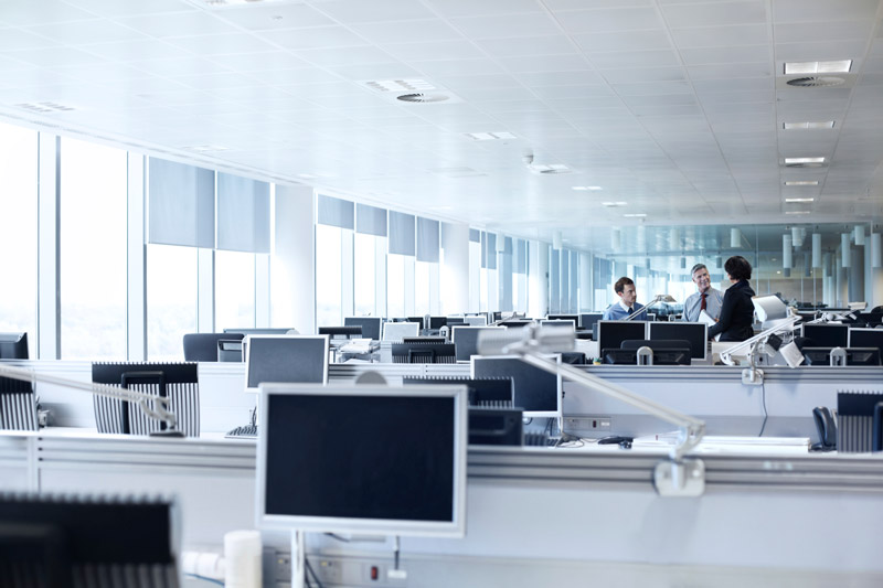 Open-plan office with rows of desks and computer monitors; a small group of three coworkers chatting at the far end.