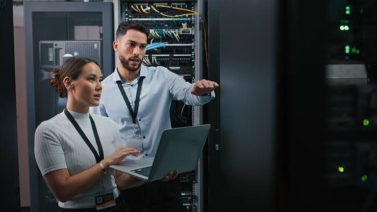 Two IT professionals in a data center review a laptop beside server racks, with one gesturing toward the equipment.