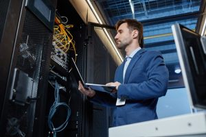 IT professional in a blue suit inspecting server racks with a laptop in a data center
