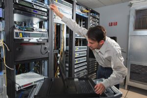 Techniker in einem Rechenzentrum greift nach Rack-Servern, mit einem Laptop auf einem Rollwagen in der Nähe und Netzkabeln in den Racks.