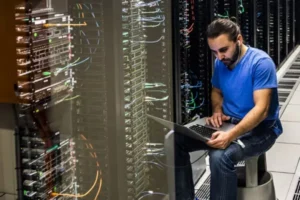 Tech-Mitarbeiter in einem blauen Hemd sitzt neben Server-Racks und benutzt in einem Rechenzentrum einen Laptop zur Fehlerbehebung.