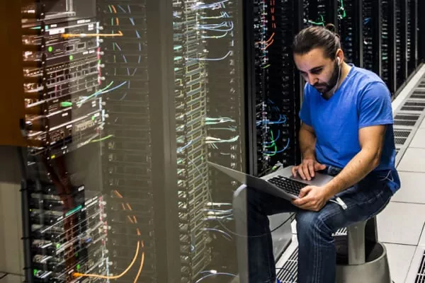 Tech worker in a blue shirt sits beside server racks, using a laptop in a data center to troubleshoot.